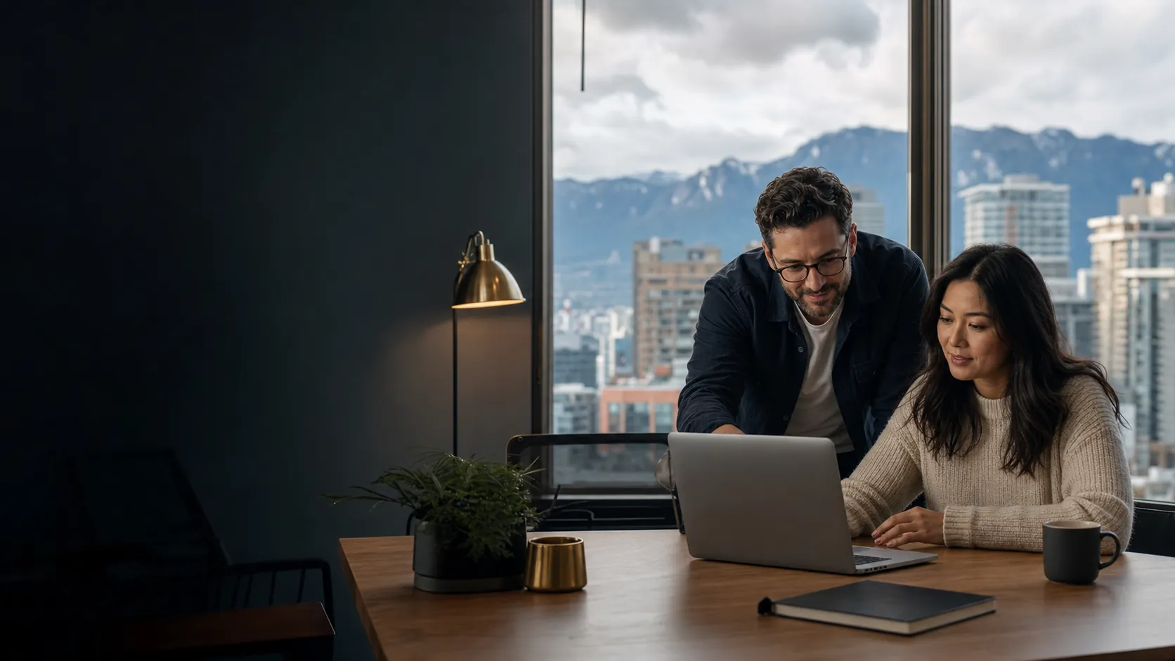 An IT consultant and a client reviewing work on a laptop, with the Vancouver skyline and mountains through the window.